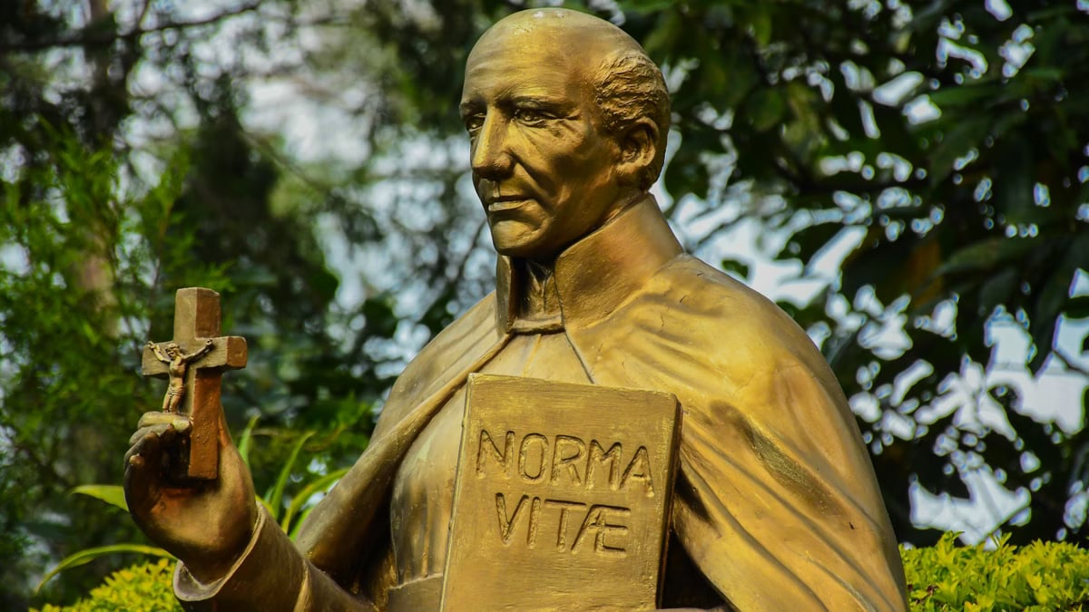 A bronze statue of a religious figure holding a cross and a book outdoors.